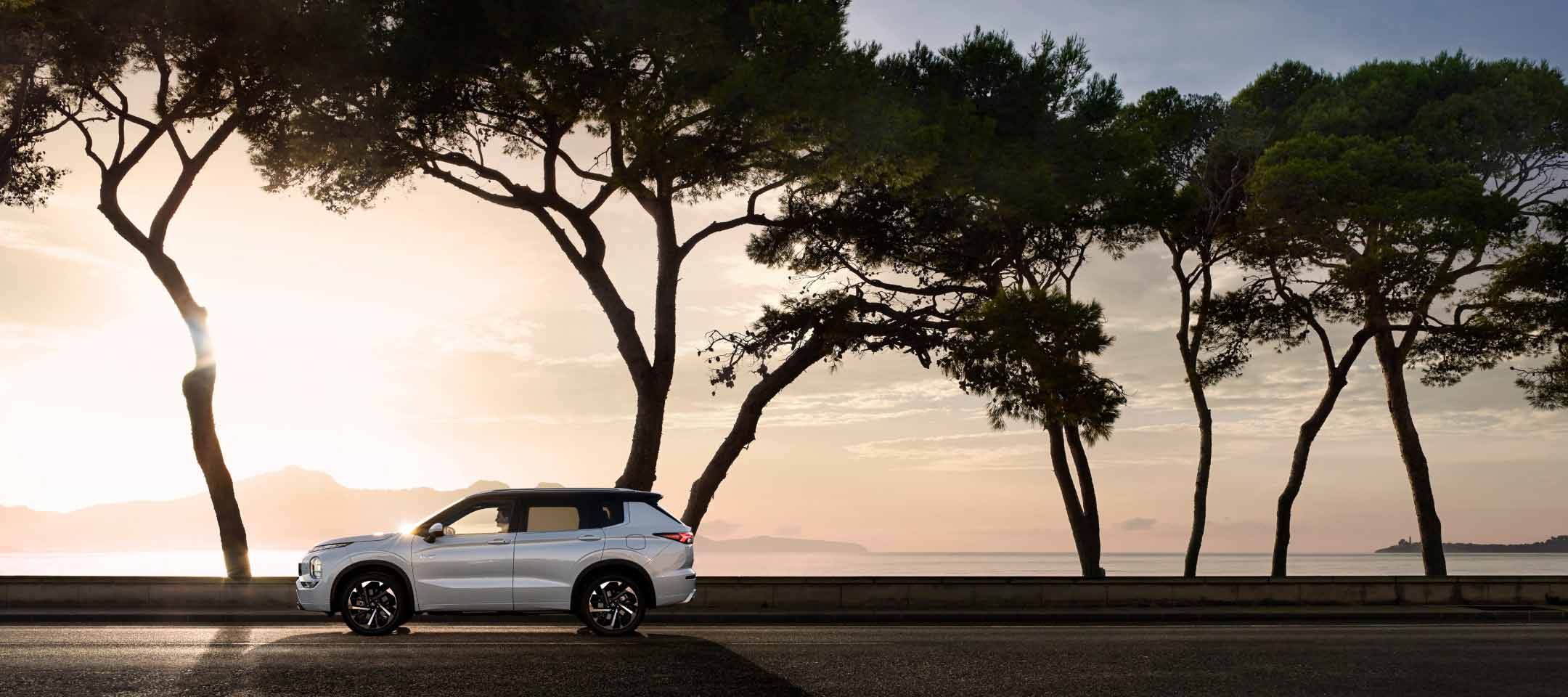 Side profile of a silver 2023 Mitsubishi Outlander PHEV SUV driving on a road with trees