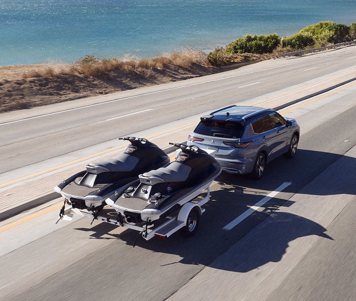 2026 Mitsubishi Outlander SUV driving on a highway beside water and towing two personal watercrafts while engaging the Trailer Stability Assist system.