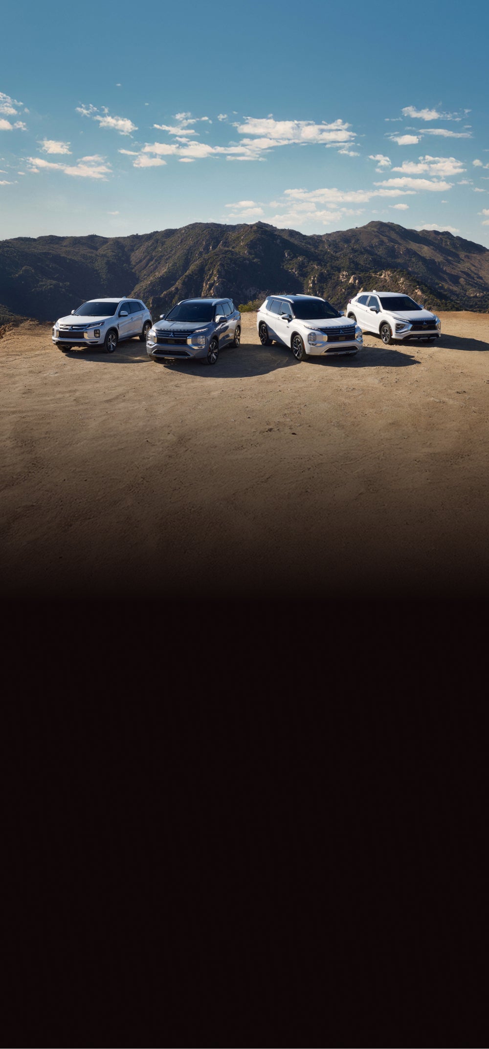 A four vehicle lineup of Mitsubishi vehicles parked in front of a mountain range. 