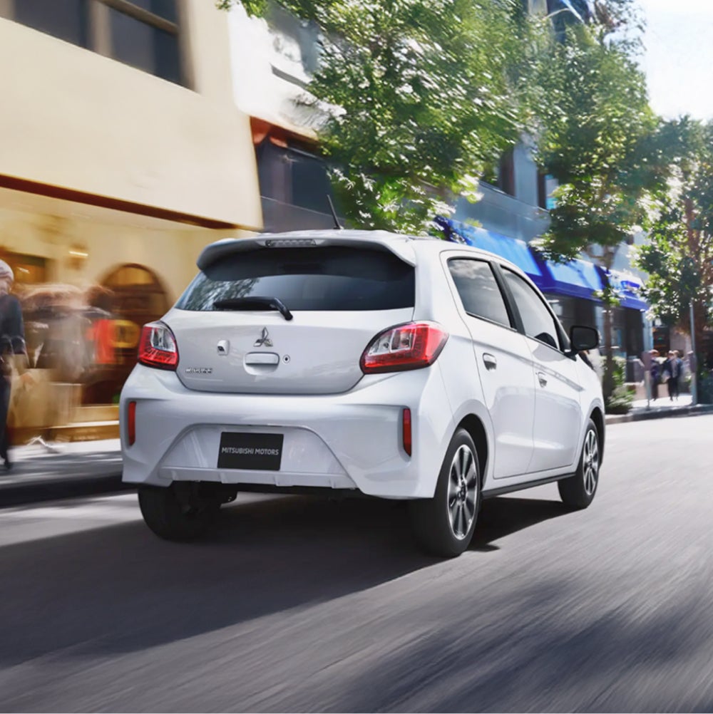 View from behind of a white Mitsubishi Mirage driving past shops on a street. 