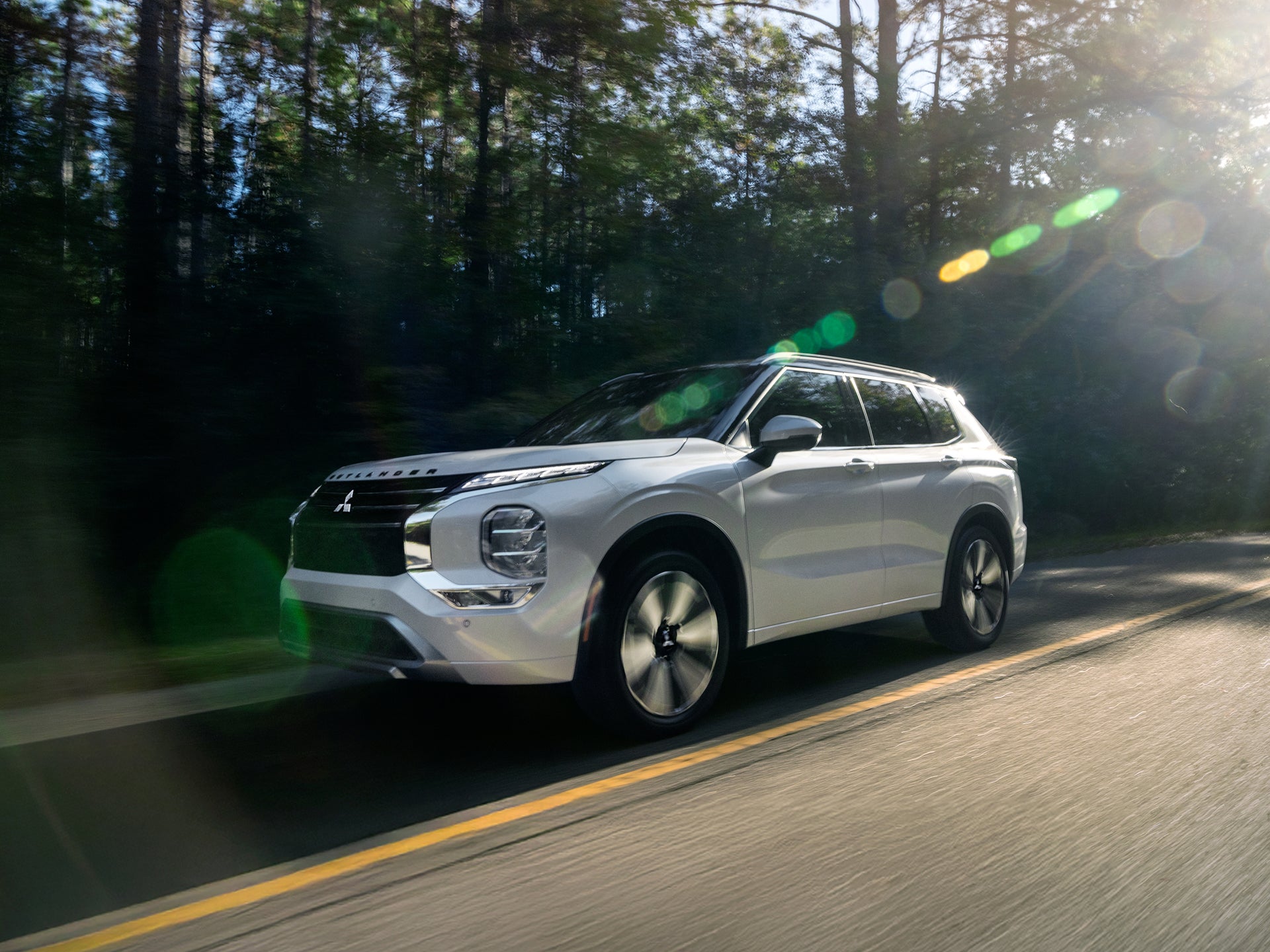Angled driver side view of a white 7 seater Mitsubishi Outlander SUV driving on a tree lined street.