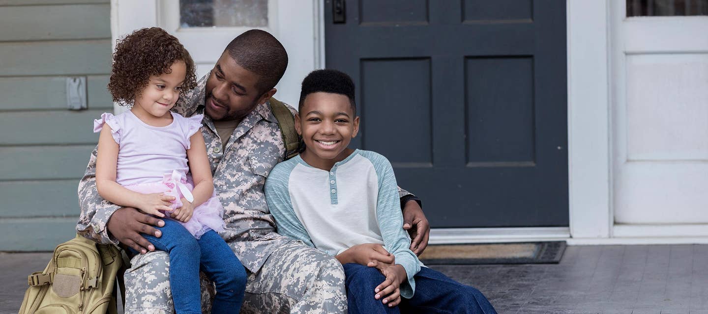 A uniformed soldier with his two small children sitting next to him on a porch outside their home