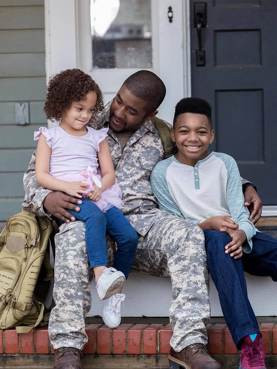 A uniformed soldier with his two small children sitting next to him on a porch outside their home